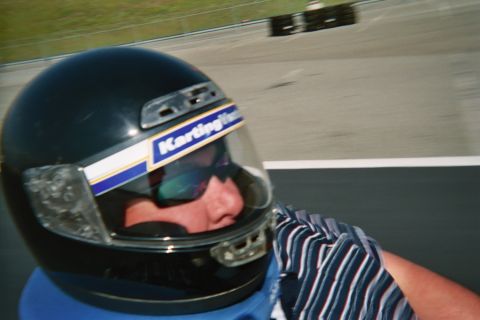 1990 Ford Bronco - Side selfie of a driver in a dark Karting Ventures helmet and sunglasses while another kart races on the track behind.