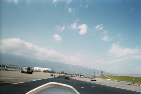 1990 Ford Bronco - Forward view over a go-kart's nose down a wide paved straight toward red-and-white barriers, buildings and mountains under blue sky.