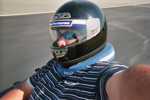 1990 Ford Bronco - man in a dark full-face racing helmet, mirrored sunglasses and a striped sleeveless shirt takes a selfie while on a kart track.