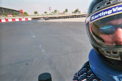 1990 Ford Bronco - Motion-blurred in-kart selfie with a dark Karting Ventures helmet at frame edge and the gray track surface streaking past.