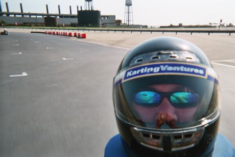 1990 Ford Bronco - Close selfie of a driver in a dark Karting Ventures helmet and mirrored sunglasses, with the open track, barriers and water towers behind.