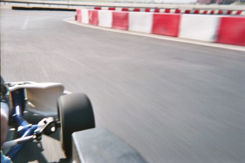 1990 Ford Bronco - Motion-blurred rear view from a go-kart as it corners past a row of red-and-white barriers on a gray paved track.