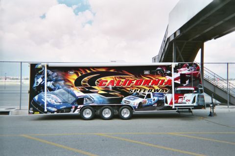 1990 Ford Bronco - large enclosed trailer wrapped with California Speedway graphics of race cars sits parked beside a chain-link fence and grandstand.