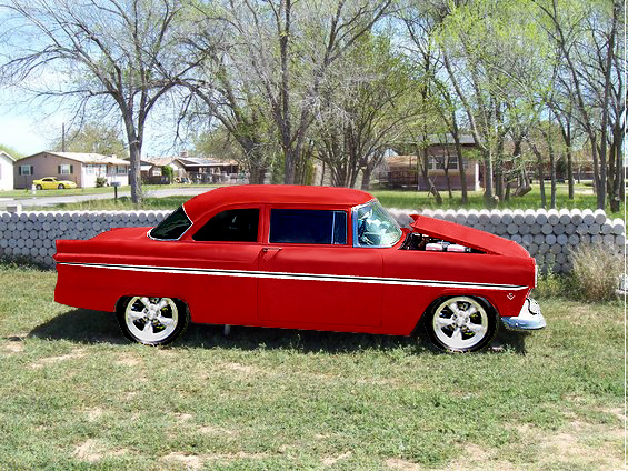 Red 1955 Ford Customline sedan parked on grass with hood open, showing the engine