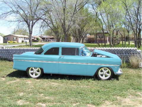 1955 Ford Customline - A blue 1955 Ford Customline sedan is parked on a grassy area with its hood open, showing the engine.