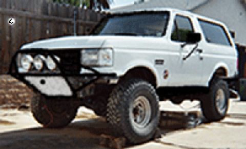 1990 Ford Bronco - white full-size Ford Bronco with a tube brush guard, KC driving lights and oversized tires sits lifted on jack stands beside a wooden fence.