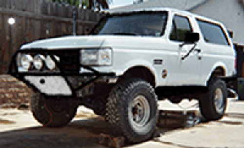 White full-size 1990s Ford Bronco with brush guard and KC lights lifted on jack stands by a wood fence