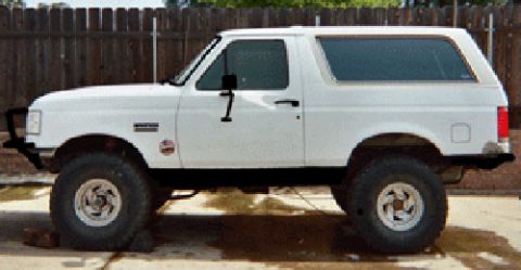 1990 Ford Bronco - Driver-side profile of a white full-size Ford Bronco, lifted on chrome wheels and mud tires, parked on concrete in front of a wooden fence.