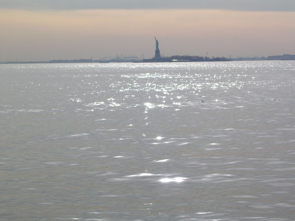 Water with sun glare, Statue of Liberty, and distant city skyline under overcast sky
