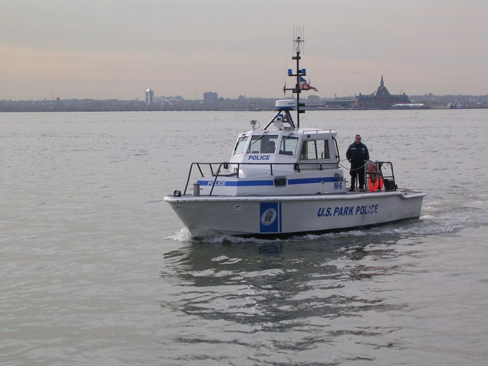 U.S. Park Police boat with an officer on deck, moving through the water with a city skyline in the background