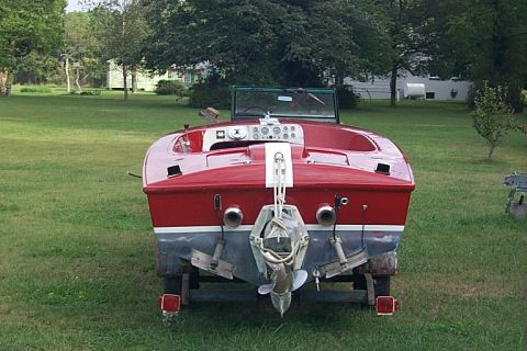 1978 Ford F-250 - A red and white boat is parked on a trailer in a grassy yard, showing its rear end with dual exhaust pipes and a propeller.