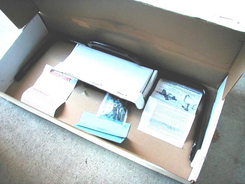 Open box with black steel suspension parts, hardware bag, and installation instructions on concrete.