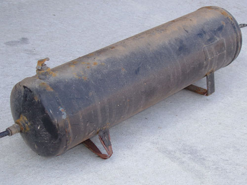 Rusty black cylindrical steel air tank with welded mounting feet on concrete