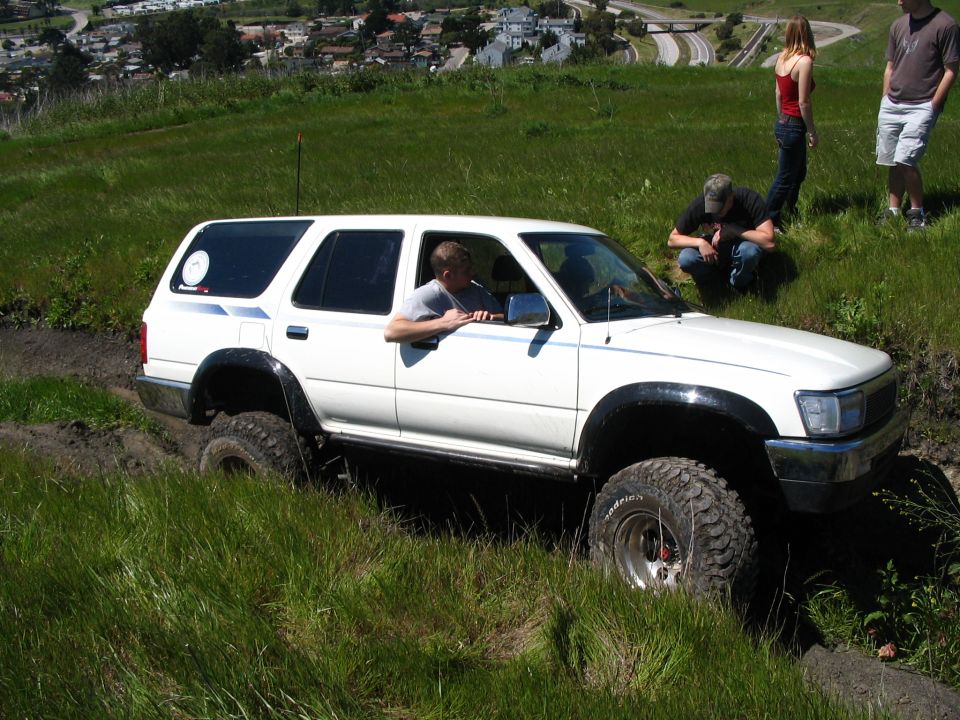 White lifted 4Runner crawling down a grassy slope on a mud track, spectators above, city and hills behind