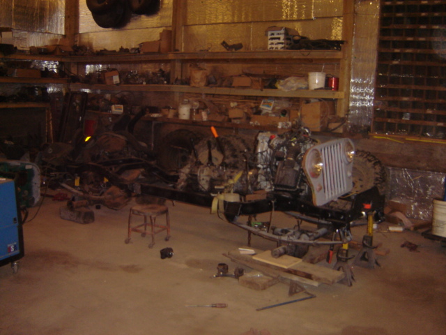 Jeep CJ frame with engine and grille assembly in a workshop. The vehicle is on jack stands