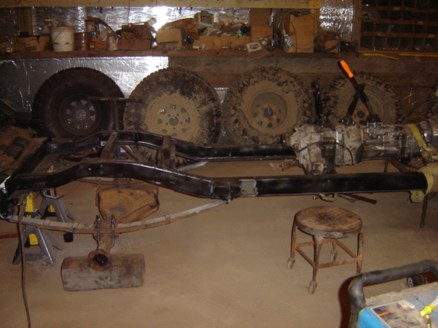 Vehicle frame on jack stands with transmission and transfer case attached, plus spare tires in background