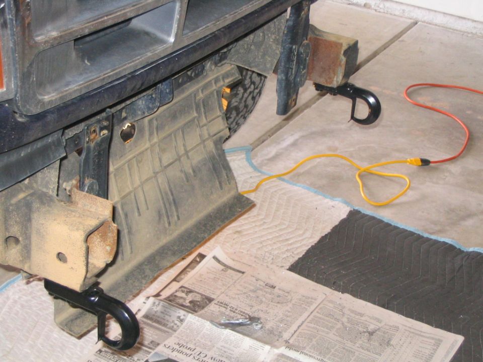 Front underside of a dark Bronco showing black tow hooks on rusty frame brackets in a garage