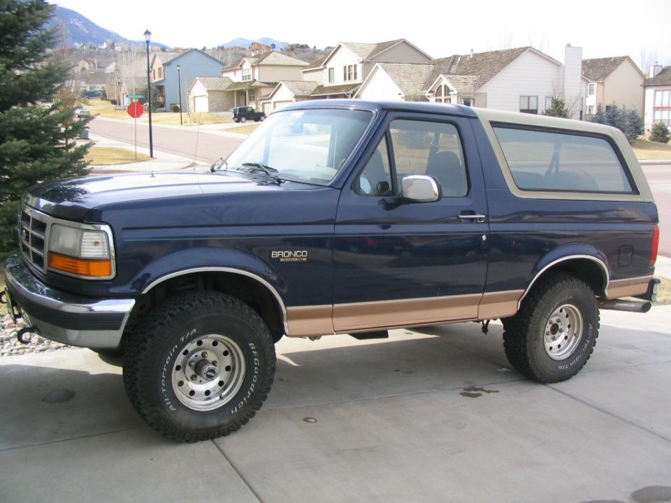 Blue and tan two-door Ford Bronco, front three-quarter view, lifted on chrome wheels in a driveway