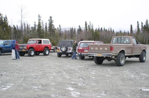 2003 Jeep TJ - Multiple off-road vehicles are parked in a gravel lot with a forest in the background.