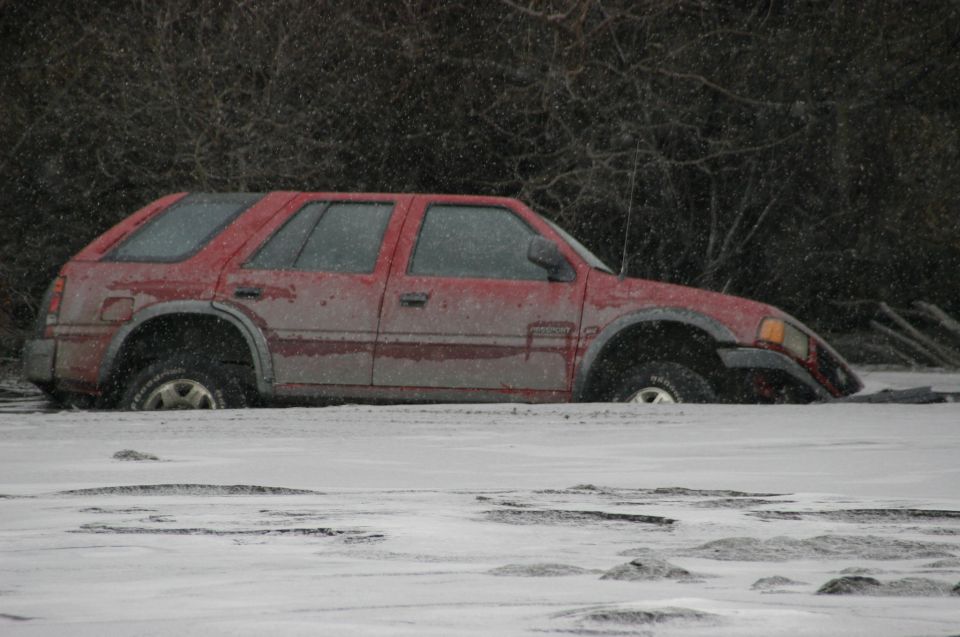 Red SUV partially submerged in snow, side view, with trees in background