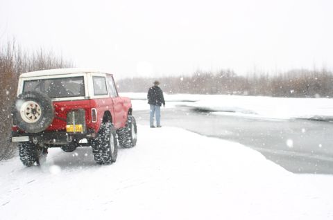 2003 Jeep TJ - A person stands by a frozen river next to a red Ford Bronco during a snowstorm.