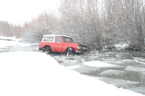 2003 Jeep TJ - A red 1978 Ford Bronco navigates through a partially frozen body of water amidst falling snow and bare trees.
