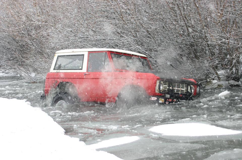 Red classic Ford Bronco driving through icy water with snow on the banks