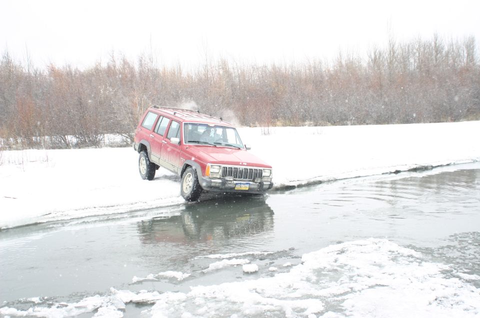 Red Jeep Cherokee XJ driving through shallow icy water with snow on the banks