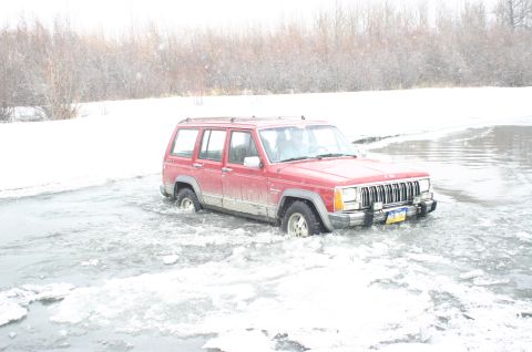 2003 Jeep TJ - A red Jeep Cherokee XJ navigates through shallow, icy water with snow-covered banks in the background.