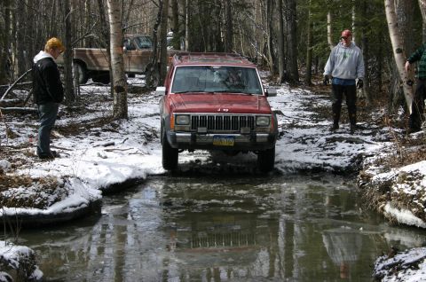 2003 Jeep TJ - A red Jeep Cherokee XJ navigates a snowy, icy stream with two people watching.