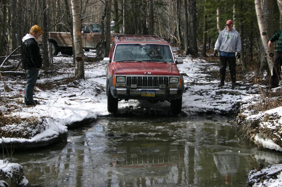 Red Jeep Cherokee XJ driving through icy water, front view. License plate is visible