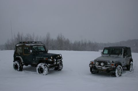 2003 Jeep TJ - Two Jeep Wranglers navigate a snowy landscape under overcast skies, with a line of trees visible in the distance.