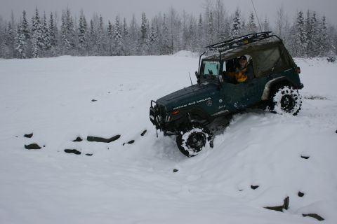 2003 Jeep TJ - A green Jeep Wrangler navigates a snowy terrain, equipped with a roof rack and large off-road tires.