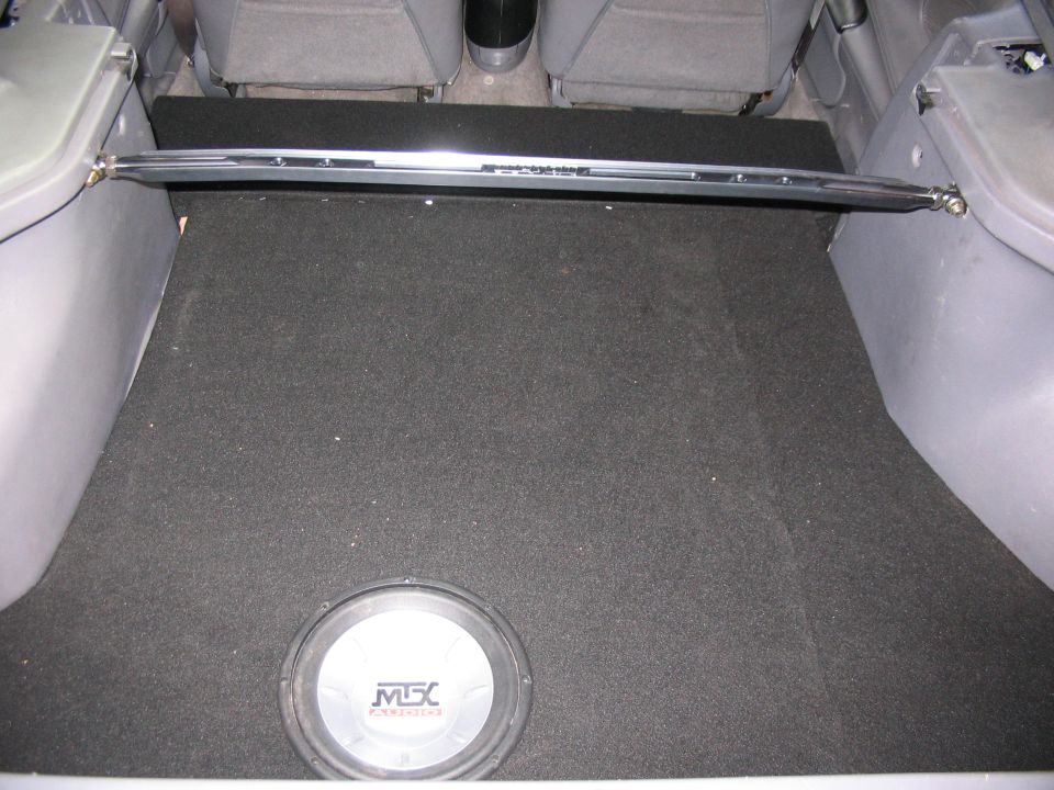 Car trunk interior showing a subwoofer and a chrome strut bar above the carpeted floor