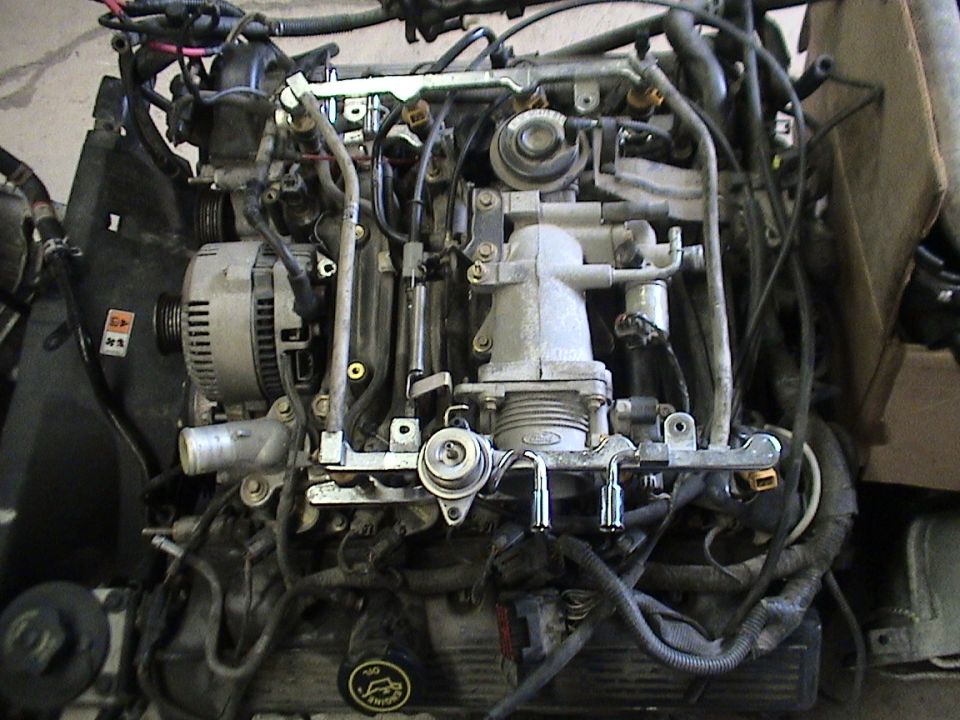 Front of a removed pushrod V-engine showing thermostat housing, fuel rail, alternator and wiring