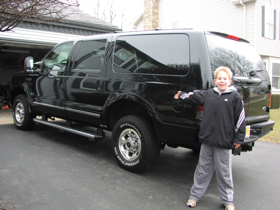 Black Ford Excursion SUV in a driveway with a boy posing beside its rear, gesturing at the vehicle