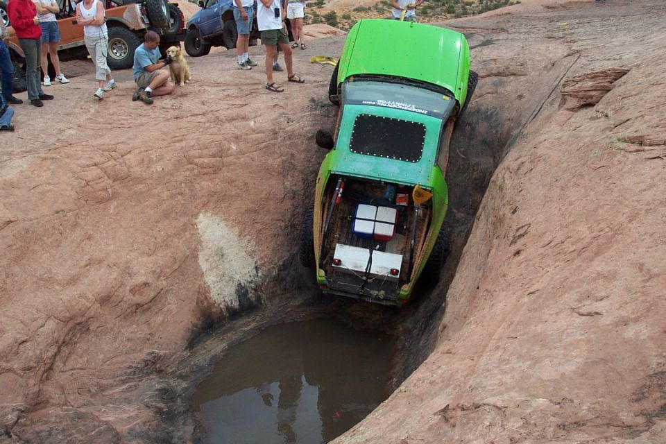Green rock crawler nosed into a water-filled slickrock hole with spectators above