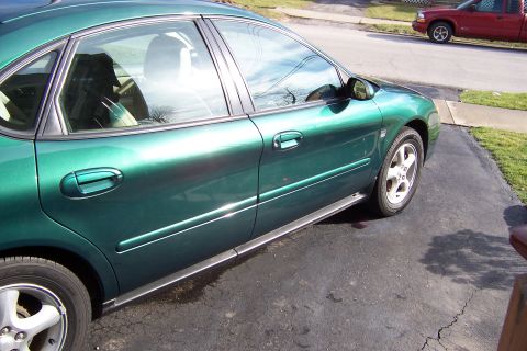 2000 Ford Taurus - A green 2000 Ford Taurus sedan is parked on a driveway, with a red pickup truck visible on the street.