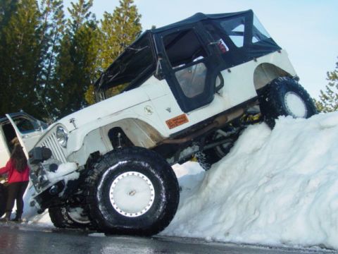 1986 Ford Bronco - A white Jeep Wrangler is angled up a large snowdrift, with its driver's side door open and a person standing nearby.