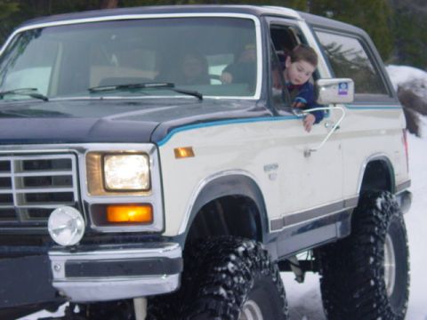 1986 Ford Bronco - A white and blue 1986 Ford Bronco with large off-road tires is seen in a snowy environment, with a child looking out the driver's window.