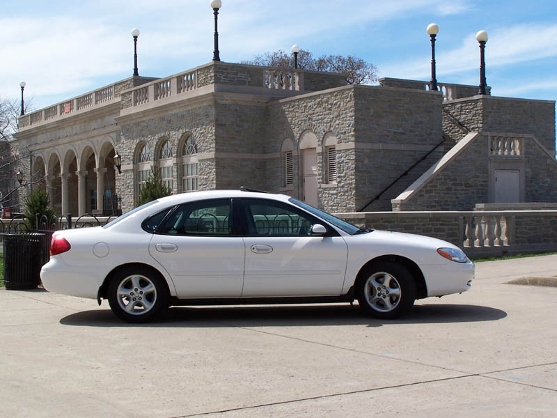 White sedan parked on concrete, side view. Building with stone facade and arched windows in background