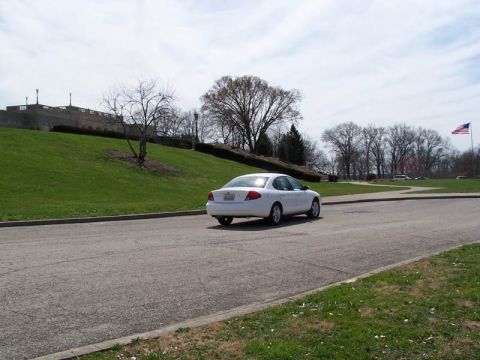 2001 Ford Taurus - A white sedan is seen from the rear driving on a paved road with a large grassy hill and trees under a cloudy sky.