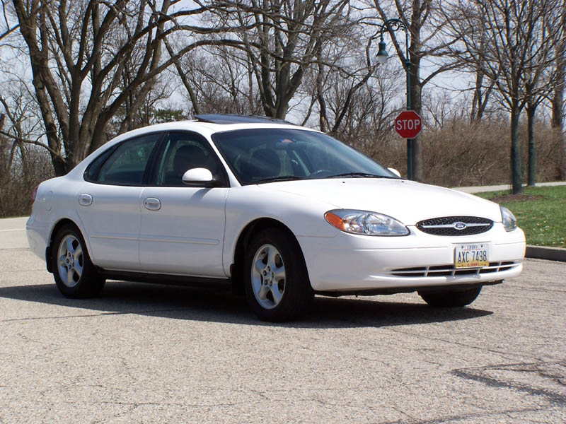 White sedan parked on asphalt, front quarter view. Visible license plate and stop sign