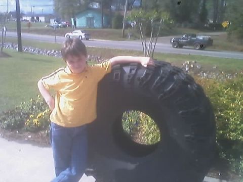 1976 Ford F-150 - A young boy poses next to a massive tire, with a road and vehicles in the background.