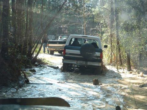 1993 Ford Bronco - Two Ford Broncos navigate a muddy, water-filled trail in a forest setting.