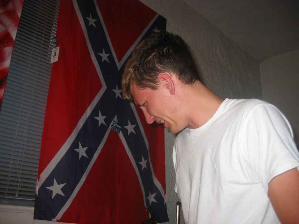 Man in white t-shirt looking down, confederate flag behind him