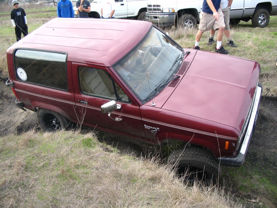 Red SUV stuck in a ditch with people watching. It looks like a Ford Bronco II