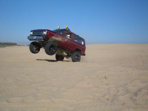 1985 Ford Bronco II - A red SUV is captured mid-air as it jumps over sand dunes under a clear blue sky.