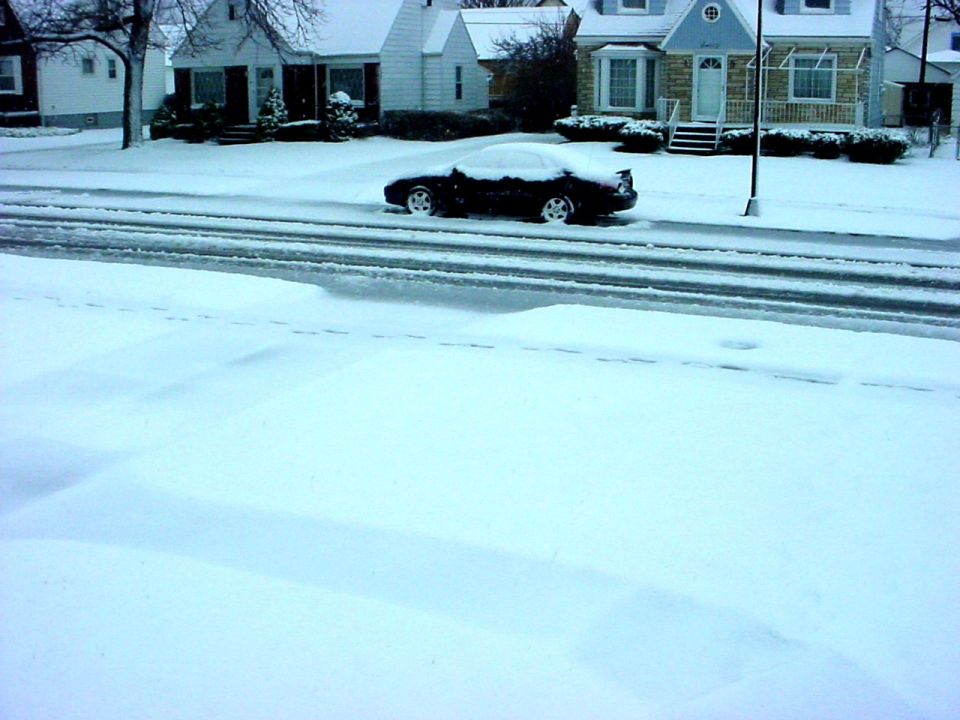 Black car covered in snow on a street with houses in the background