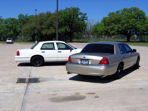 2000 Ford Crown Victoria - Two sedans, likely police cruisers, are parked side-by-side in an outdoor lot under a clear blue sky.